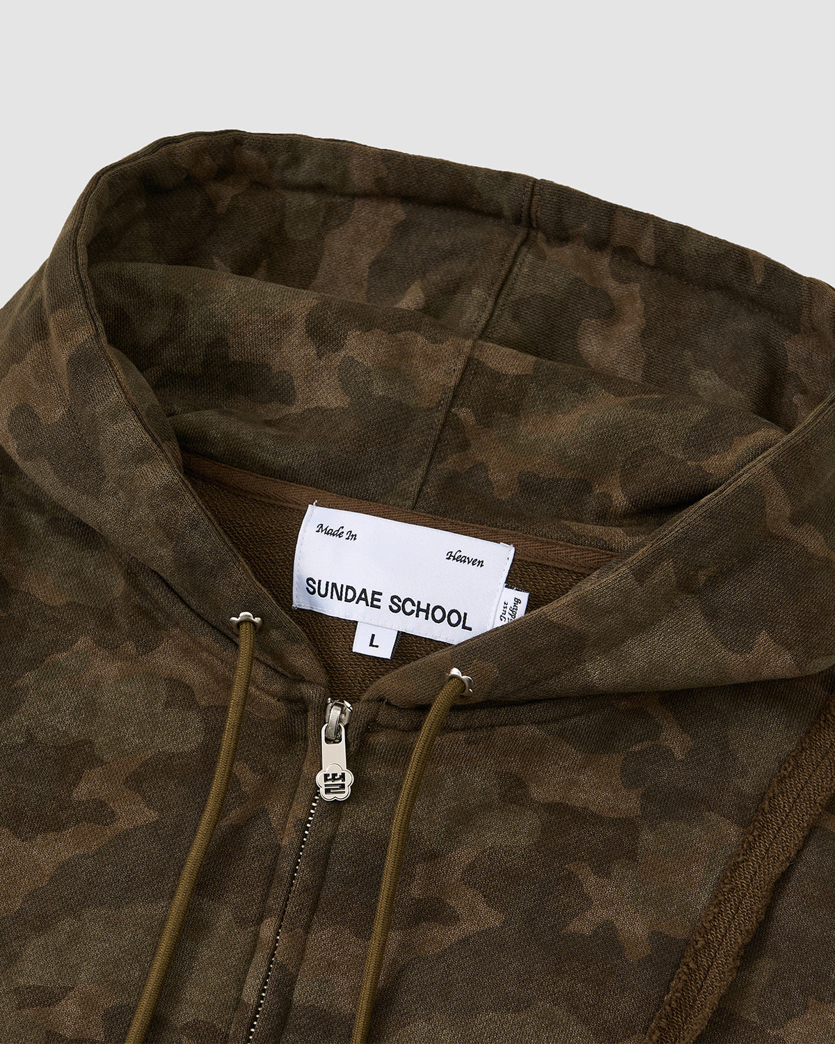 Close-up of the Sundae School CAMO MOCHI HAZE PANELED ZIP UP HOODIE in size L, featuring a camouflage pattern, zipper, and drawstrings. Made from 100% cotton in Korea. Displayed on a plain light gray background.