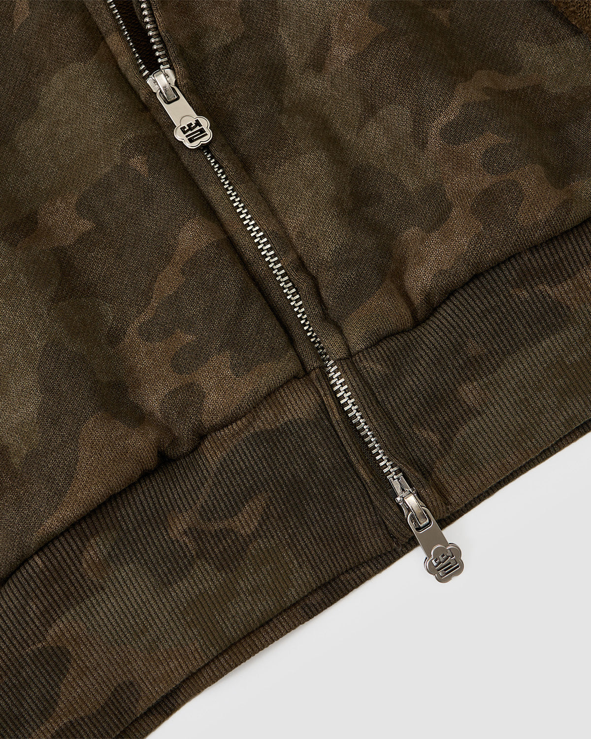 Close-up of the Sundae School CAMO MOCHI HAZE PANELED ZIP UP HOODIE, showing its green and brown camouflage panels, raw seams, metallic double zipper, and ribbed bottom edge.