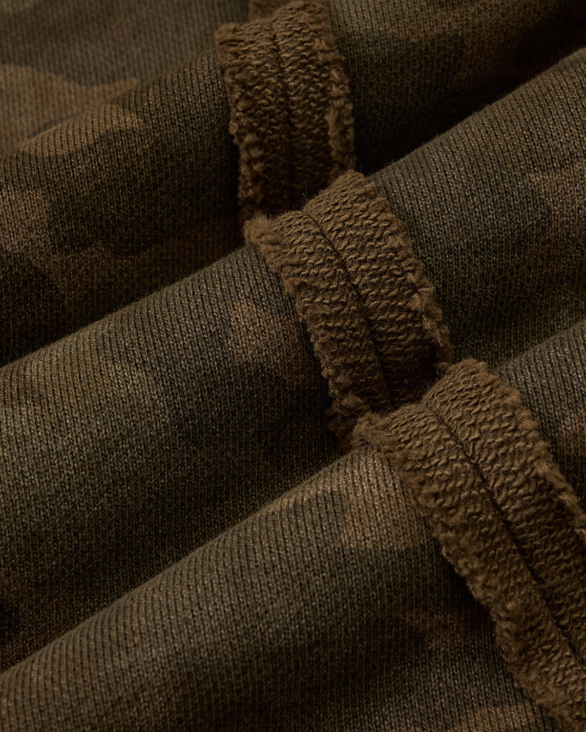 Close-up of the CAMO MOCHI HAZE PANELED ZIP UP HOODIE by Sundae School, featuring 100% cotton camouflage fabric with textured stitching and seams in green and brown tones.