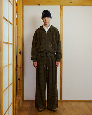 Indoors on a wooden floor, a person wears a dark camo hoodie, brown shoes, a dark beanie, and Sundae School CAMO MOCHI HAZE PANELED SWEATPANTS—wide-leg 100% cotton pants with drawstrings and raw seam panels. The space has wood paneling and white walls.