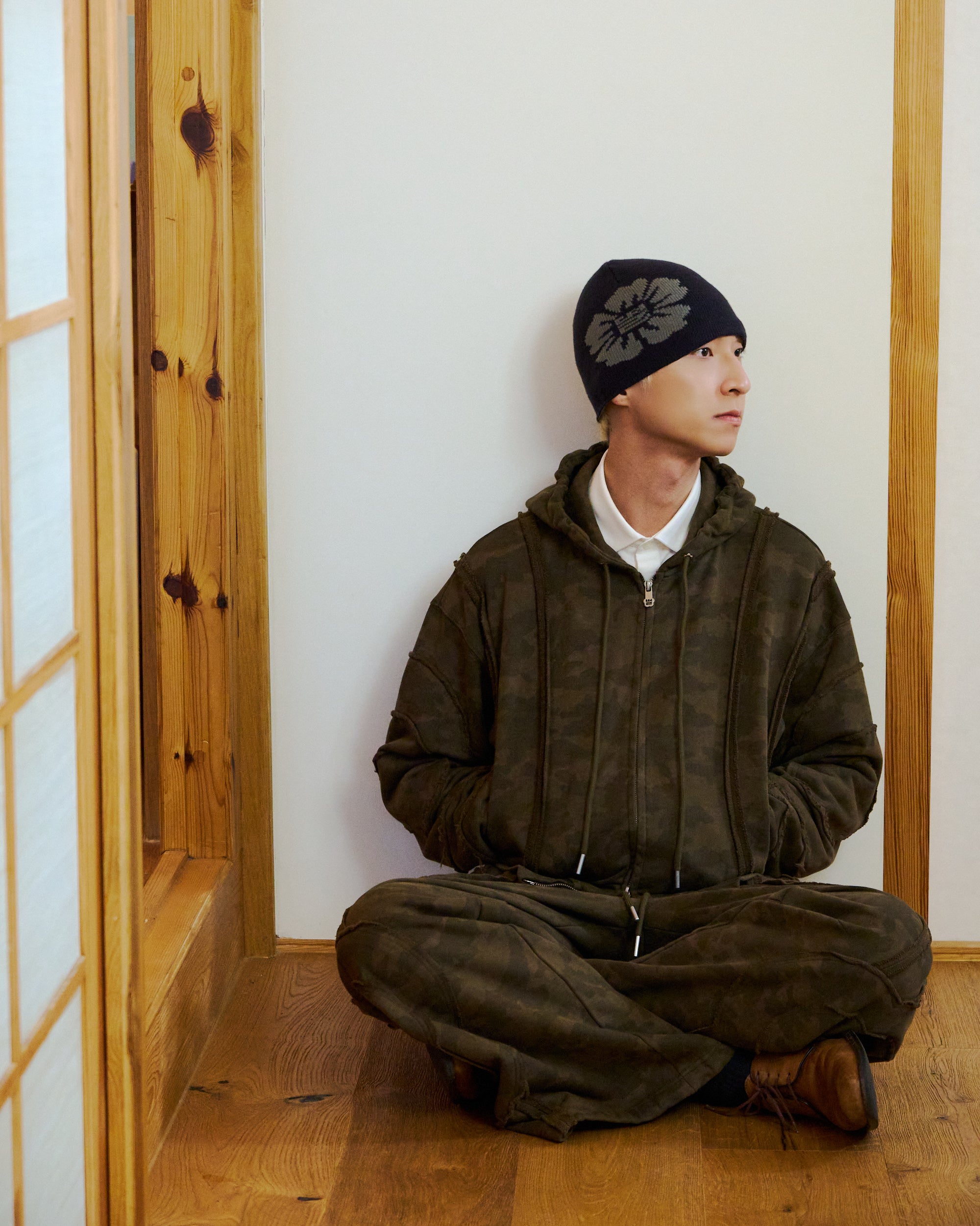 A person in Sundae School's CAMO MOCHI HAZE PANELED SWEATPANTS and a beanie with a leaf design sits cross-legged on a wooden floor, looking right, against a light-colored wall with wooden trim.