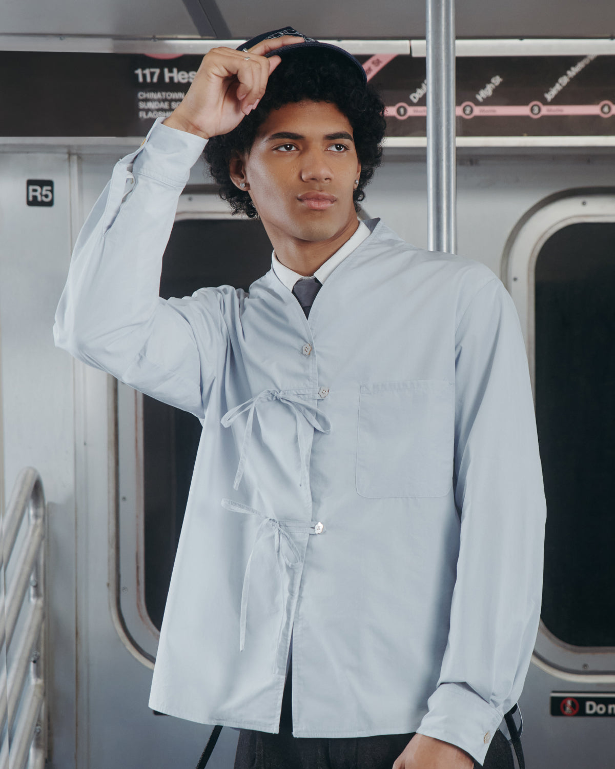 A young man wearing the Sundae School DART COLLAR SHIRT stands in a subway car, holding onto his hat. A subway map and seats are visible in the background.