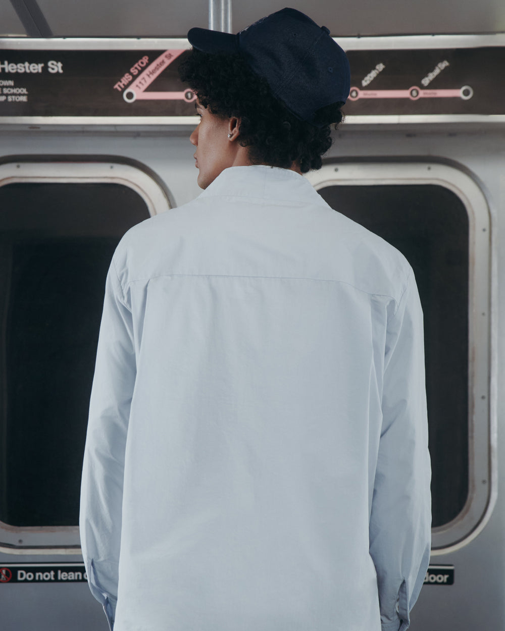 A person wearing the DART COLLAR SHIRT by Sundae School in light blue and a navy cap stands with their back to the camera on a subway, facing closed doors and a subway map above.
