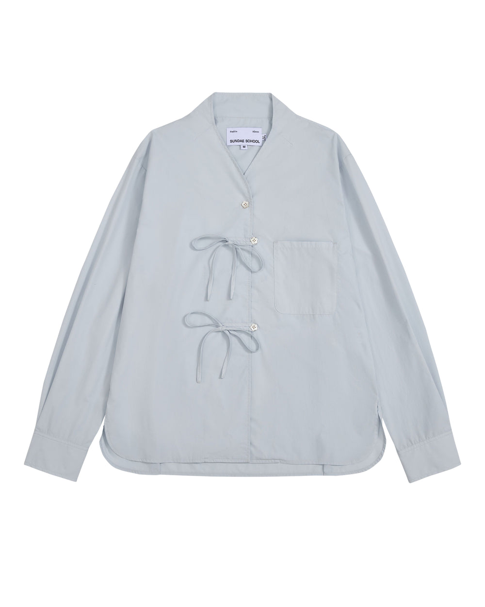 The DART COLLAR SHIRT by Sundae School is a light blue long-sleeve shirt crafted from a soft cotton-nylon blend, featuring two front tie closures, a single chest pocket, and a collarless neckline. Inspired by the relaxed silhouette of a shawl collar shirt, this men’s button-down is displayed flat against a white background.