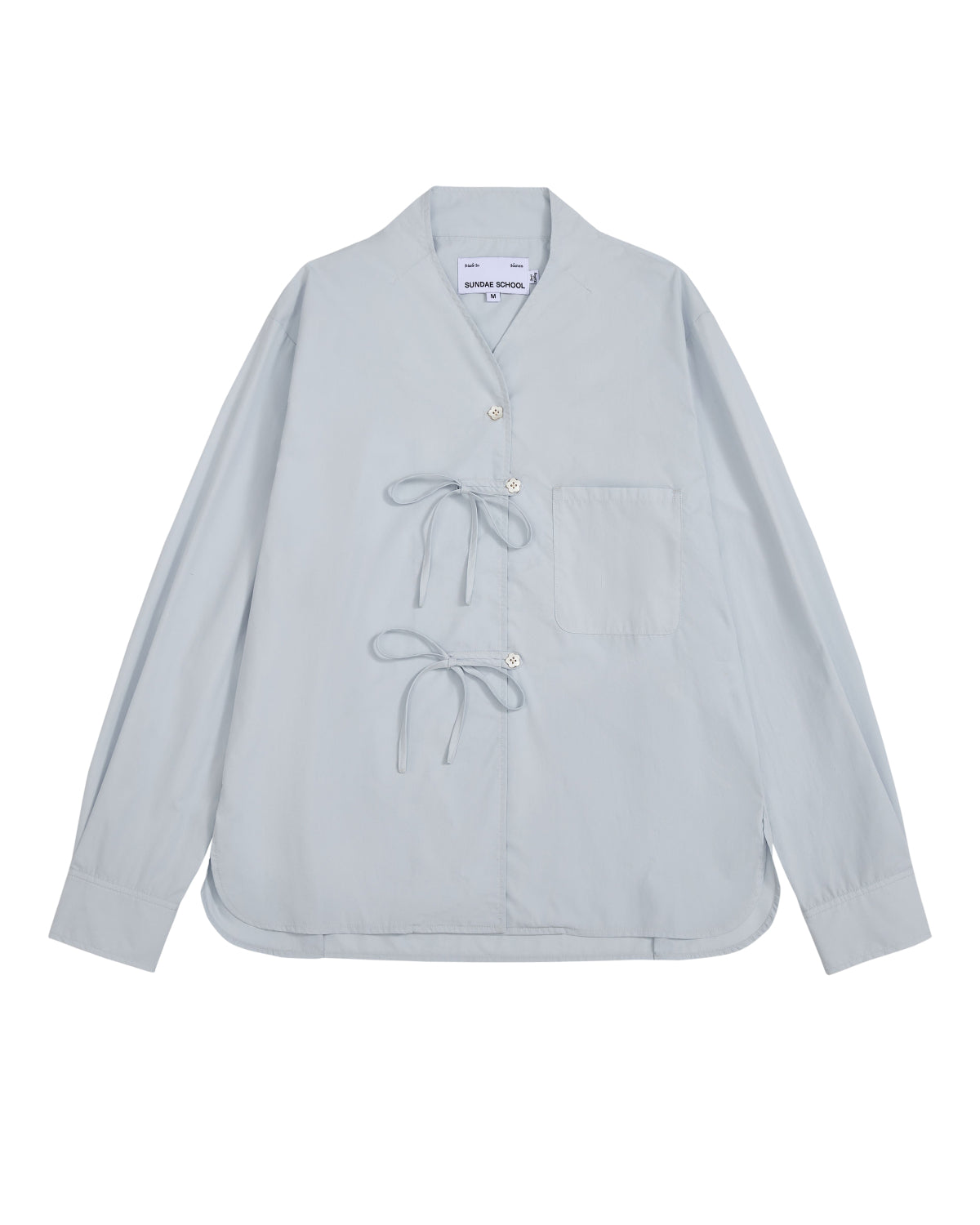 The DART COLLAR SHIRT by Sundae School is a light blue long-sleeve shirt crafted from a soft cotton-nylon blend, featuring two front tie closures, a single chest pocket, and a collarless neckline. Inspired by the relaxed silhouette of a shawl collar shirt, this men’s button-down is displayed flat against a white background.