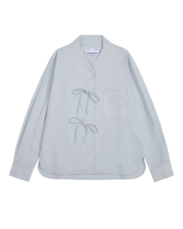 The DART COLLAR SHIRT by Sundae School is a light blue long-sleeve shirt crafted from a soft cotton-nylon blend, featuring two front tie closures, a single chest pocket, and a collarless neckline. Inspired by the relaxed silhouette of a shawl collar shirt, this men’s button-down is displayed flat against a white background.