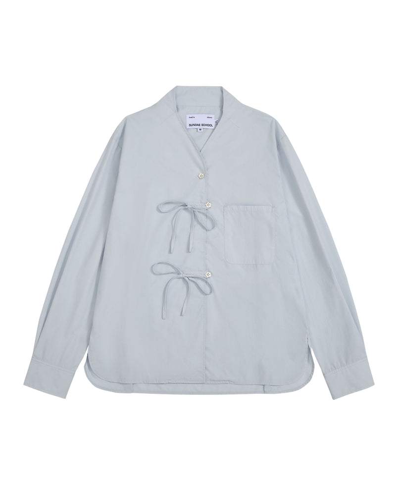 The DART COLLAR SHIRT by Sundae School is a light blue long-sleeve shirt crafted from a soft cotton-nylon blend, featuring two front tie closures, a single chest pocket, and a collarless neckline. Inspired by the relaxed silhouette of a shawl collar shirt, this men’s button-down is displayed flat against a white background.