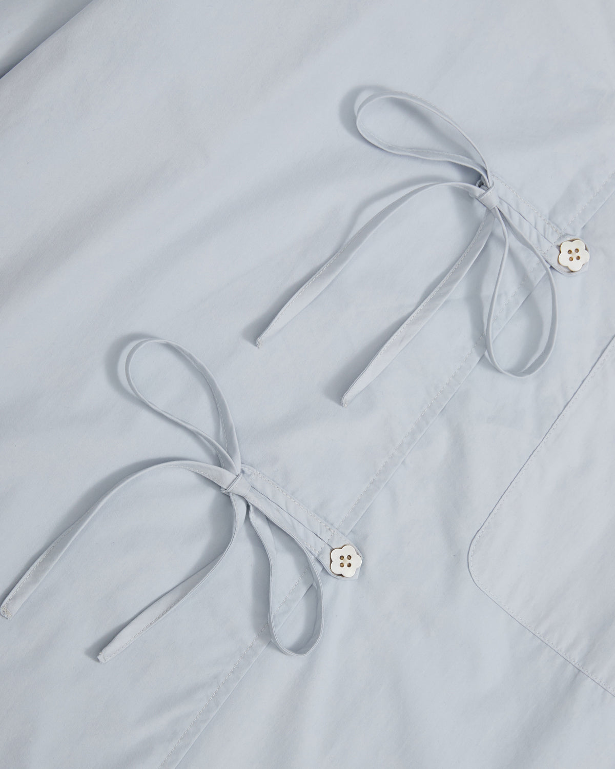 Close-up of Sundae School's DART COLLAR SHIRT in light blue fabric, featuring two tied bows and two diagonally arranged white buttons on a smooth surface.