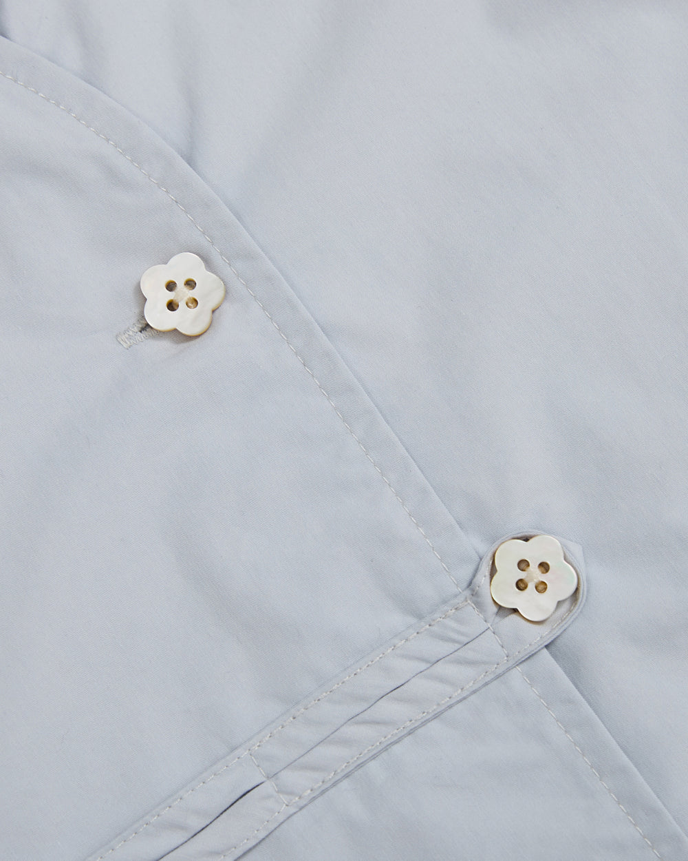 Close-up of a light blue cotton-nylon blend fabric from the Sundae School DART COLLAR SHIRT, featuring two white, four-hole flower-shaped buttons. The buttons are attached at intersecting stitched seams, adding subtle detail to this classic shirt design.
