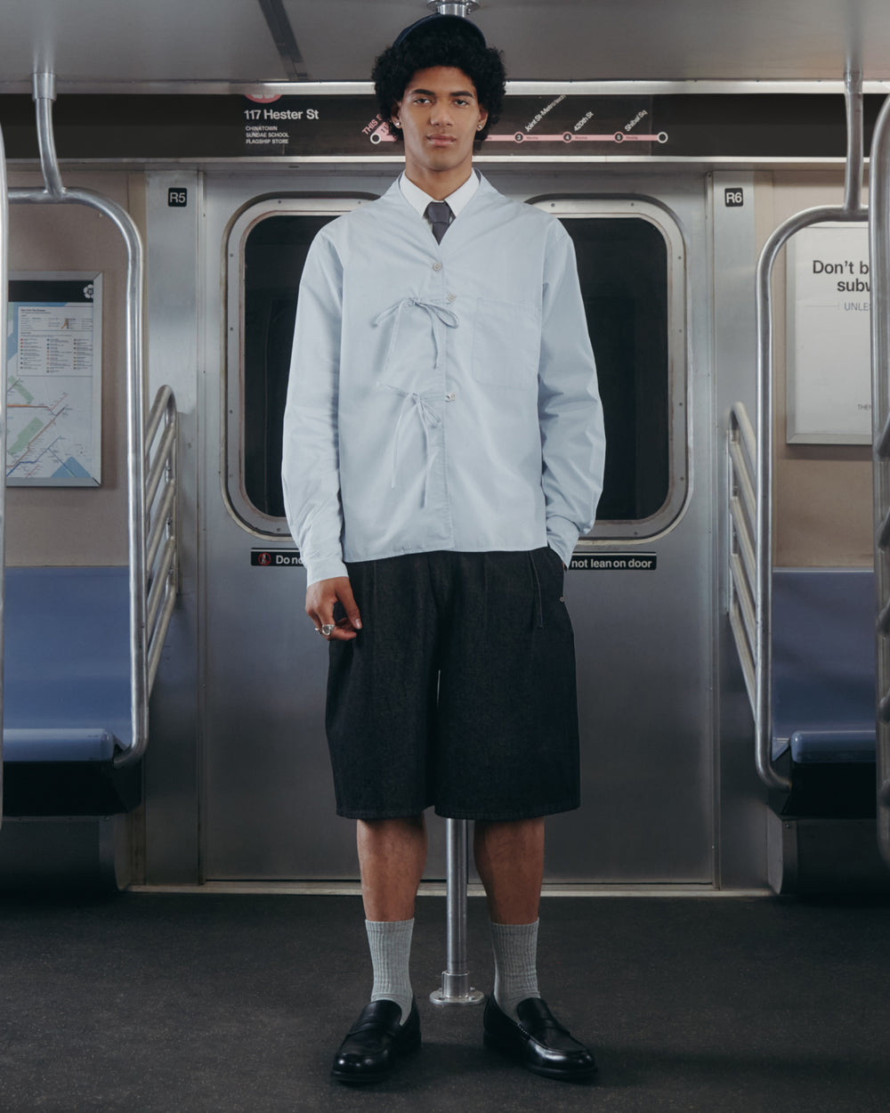 A person stands in a mostly empty subway car wearing the Sundae School DART COLLAR SHIRT, styled with dark knee-length shorts, gray socks, and black loafers.