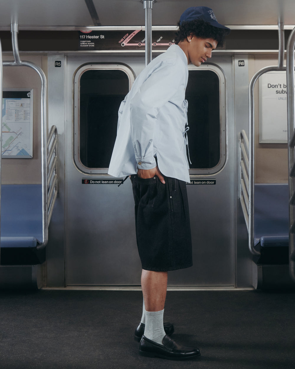 A person wearing the Sundae School DART COLLAR SHIRT, along with a blue cap, dark shorts, white socks, and black shoes, stands inside a subway train with hands in pockets and facing left. Subway seats, doors, and a route map are visible in the background.