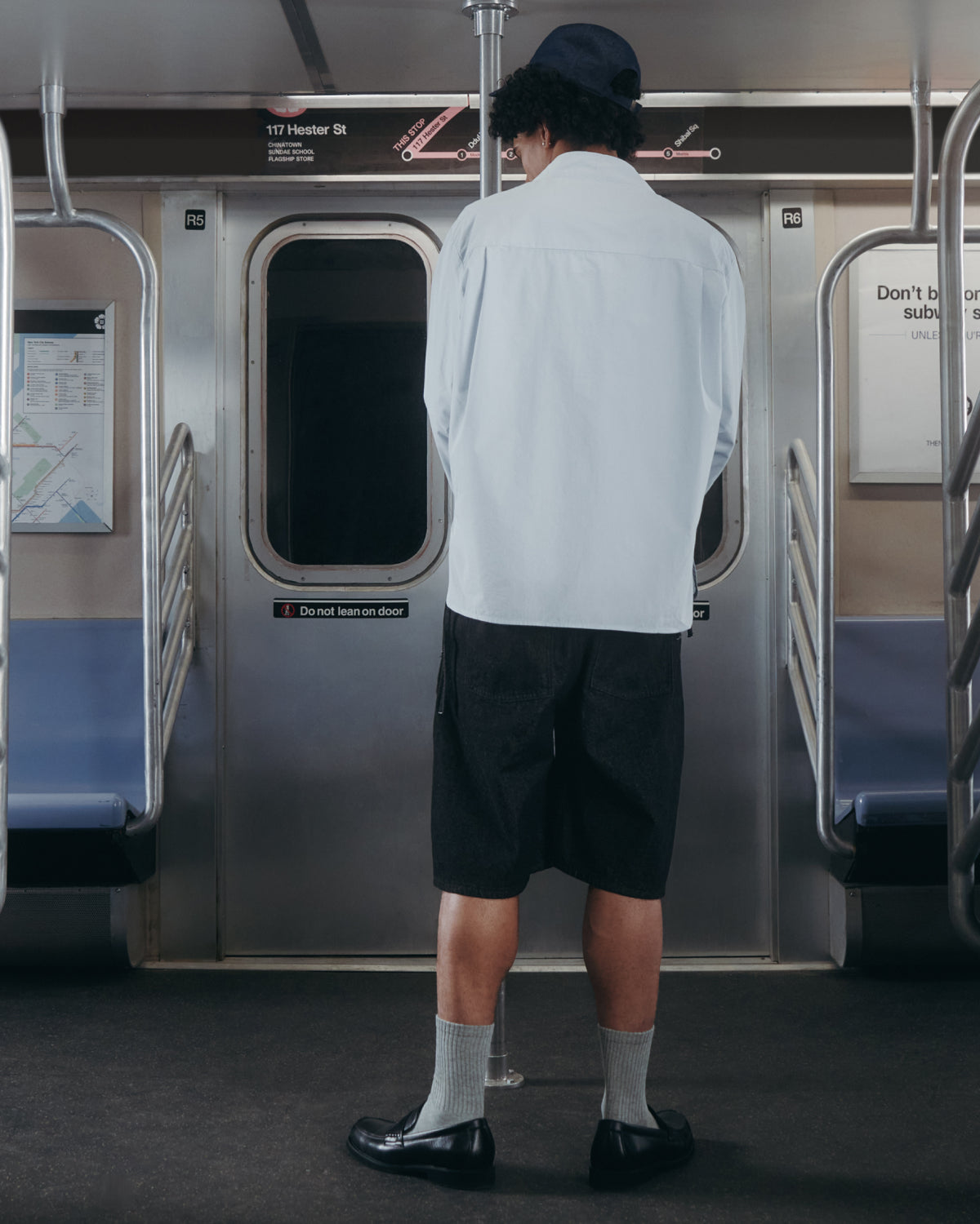 A person wearing the Sundae School DART COLLAR SHIRT, paired with black shorts and black shoes, stands facing the door inside a subway train, surrounded by empty blue seats on both sides.