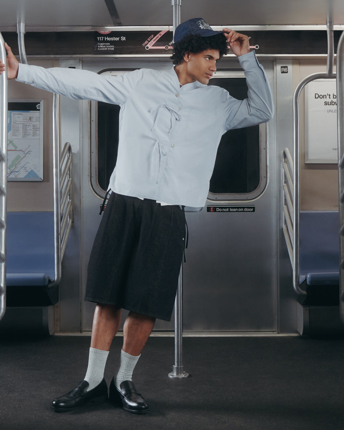 A person stylishly poses inside a subway train, holding a pole and tilting their cap while wearing Sundae School BLACK VITA JORTS, a light blue shirt, white socks, black shoes, and a blue cap.