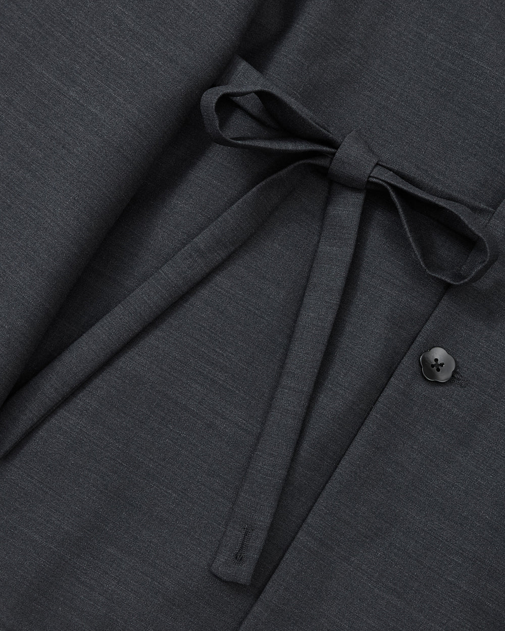 A close-up of the Sundae School CHARCOAL DOUBLE DUTY BLAZER highlights its dark gray premium wool blend, featuring a fabric belt tied in a bow for a subtle hanbok-inspired silhouette, with a single black button accenting the right side.