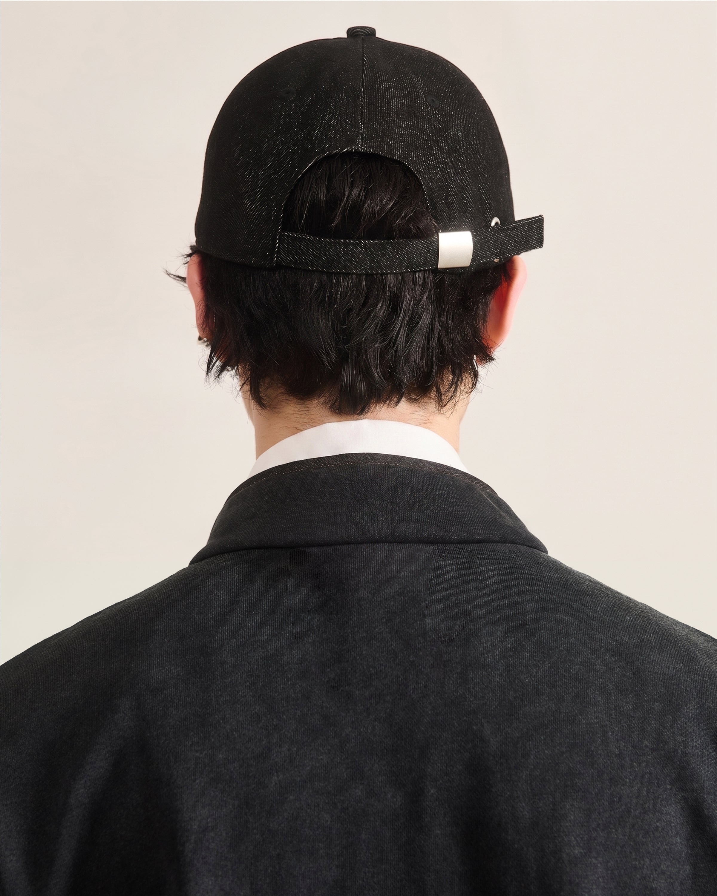 A person with short, dark hair is seen from behind, wearing a BLACK DRAGON CAP by Sundae School and a dark jacket over a white collared shirt. The background is plain and light-colored.