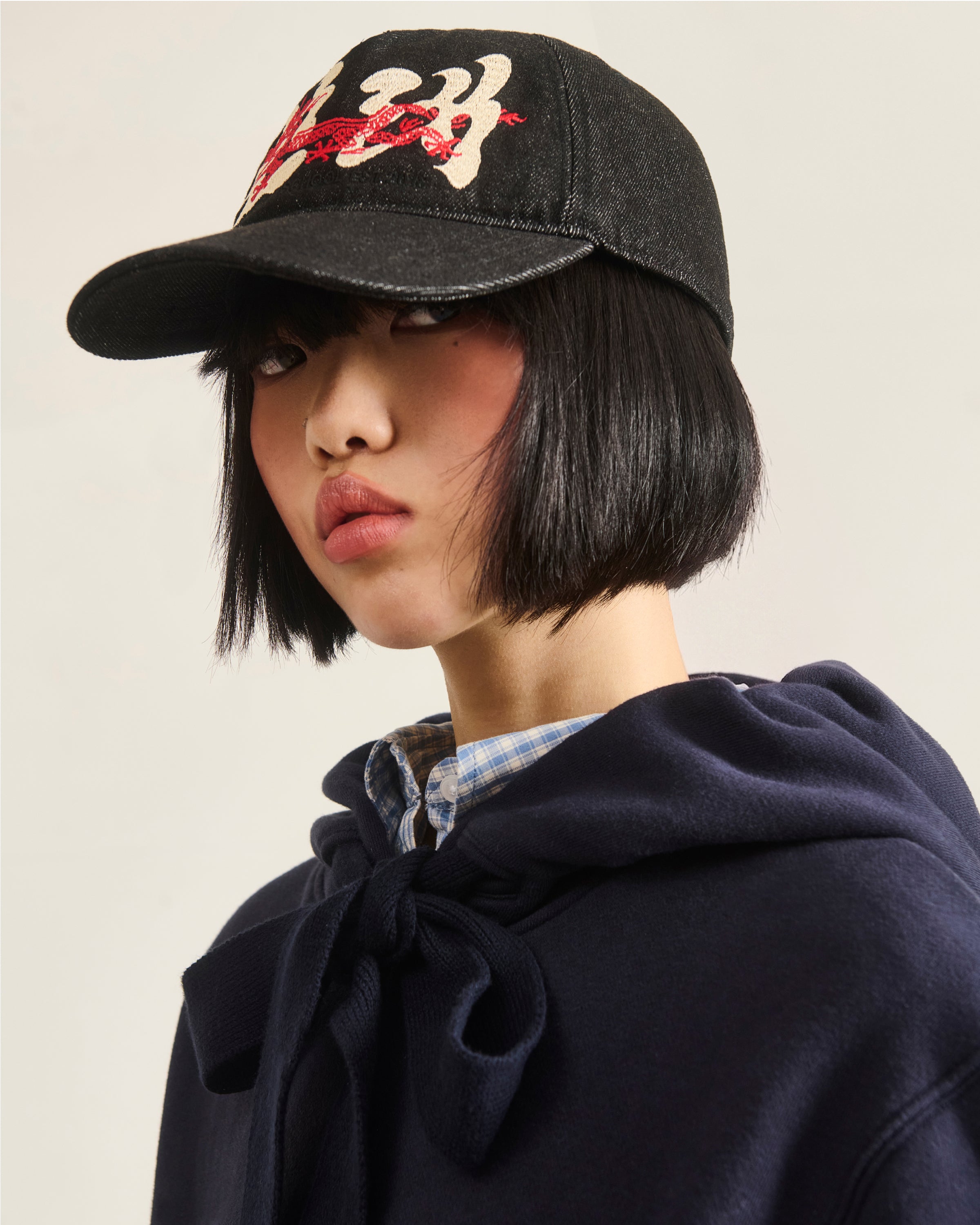 A person with a short black bob wears the Sundae School BLACK DRAGON CAP, featuring white and red embroidery, paired with a blue plaid shirt and a navy hoodie tied at the neck, gazing slightly to the side.