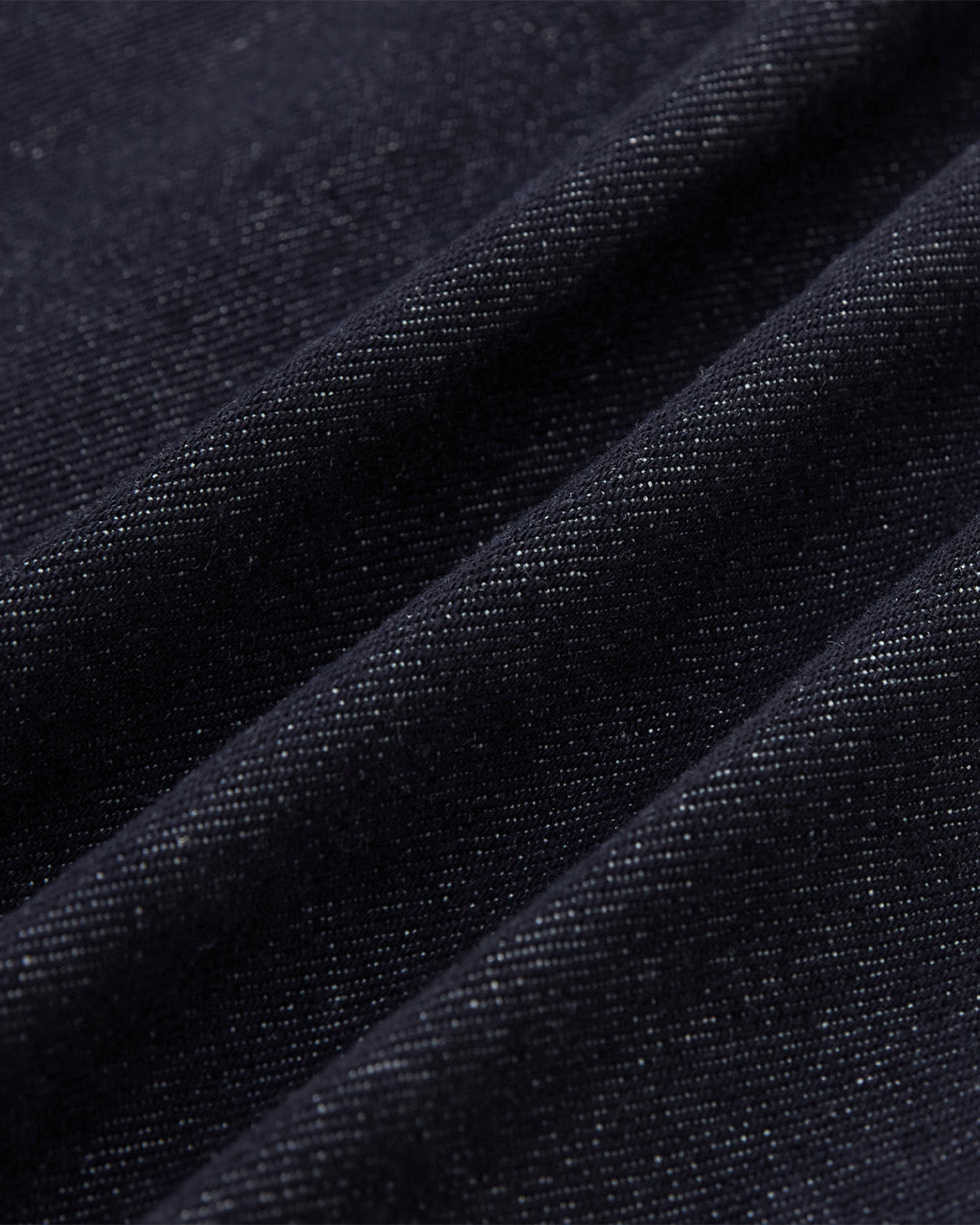 Close-up of the Sundae School DENIM CHORE JACKET in dark blue, showcasing its textured weave and subtle sheen with visible diagonal folds.