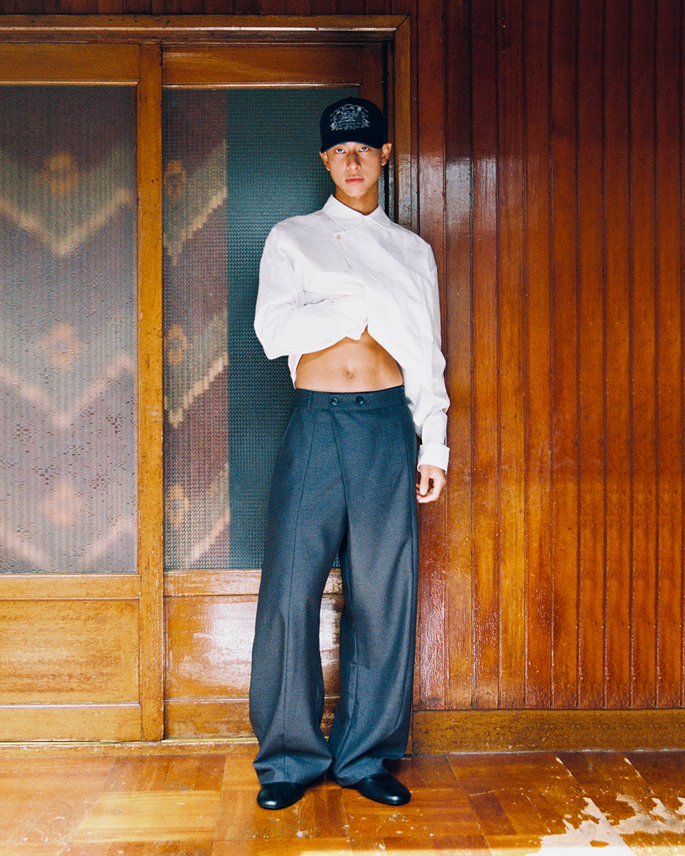 A model in a white shirt lifts it to show the waistband of Sundae School CHARCOAL PLEATED HANBOK SLACKS, standing by a wooden door.
