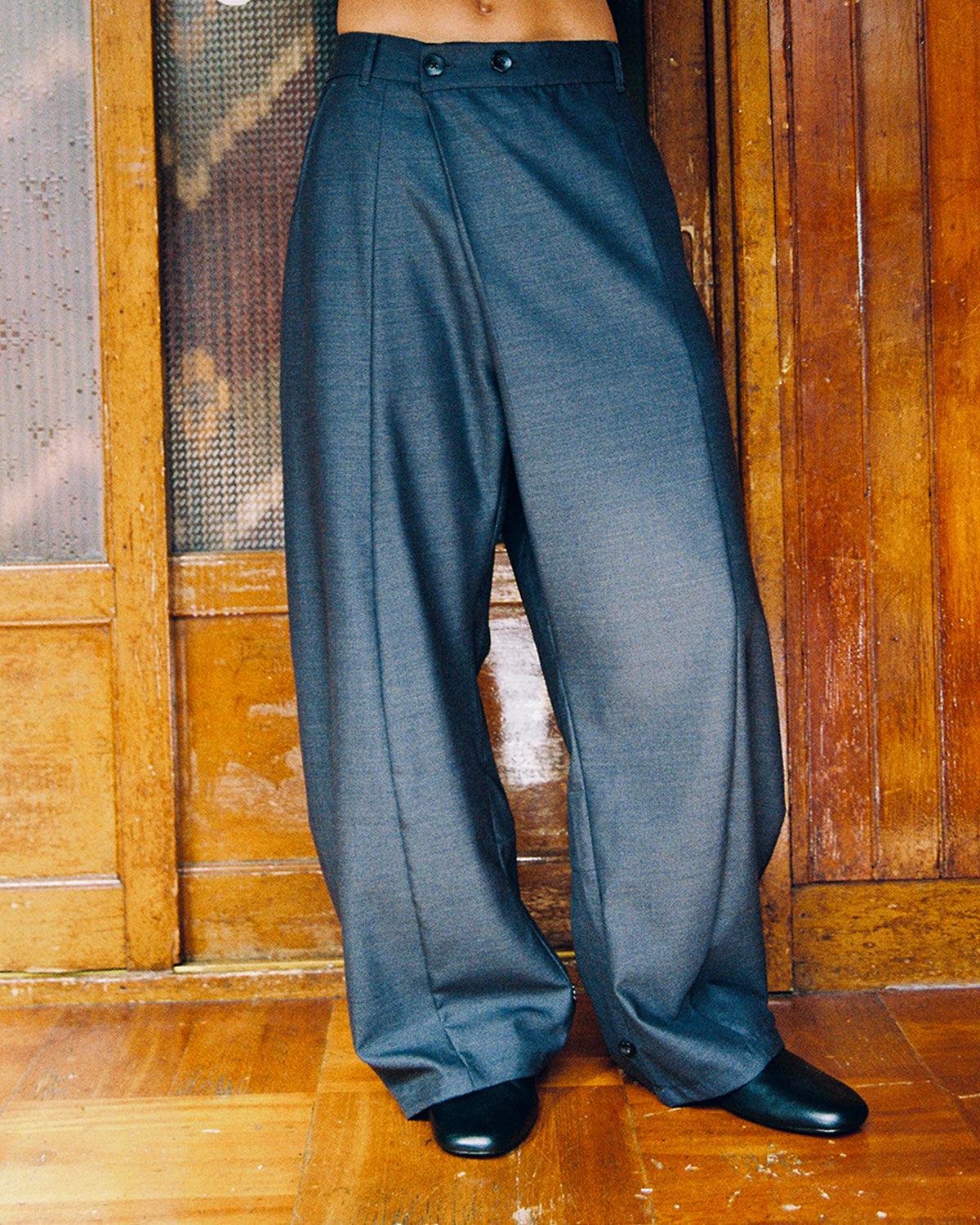 Close-up of a model wearing Sundae School CHARCOAL PLEATED HANBOK SLACKS, a wide-leg charcoal trouser with front pleats, with black shoes on a wood floor.