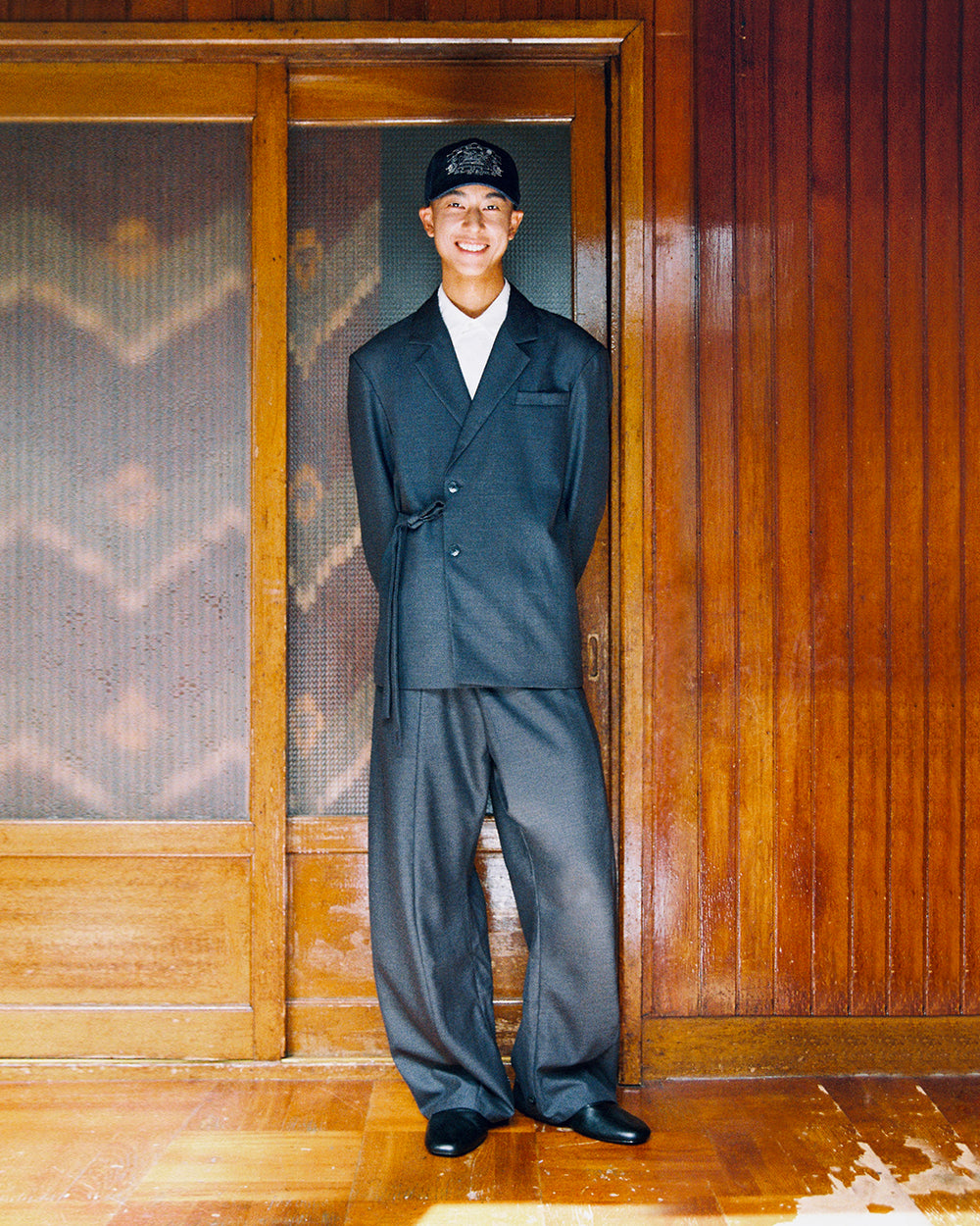 A model smiles in the Sundae School CHARCOAL DOUBLE DUTY BLAZER and matching CHARCOAL PLEATED HANBOK SLACKS, standing by a wooden door.