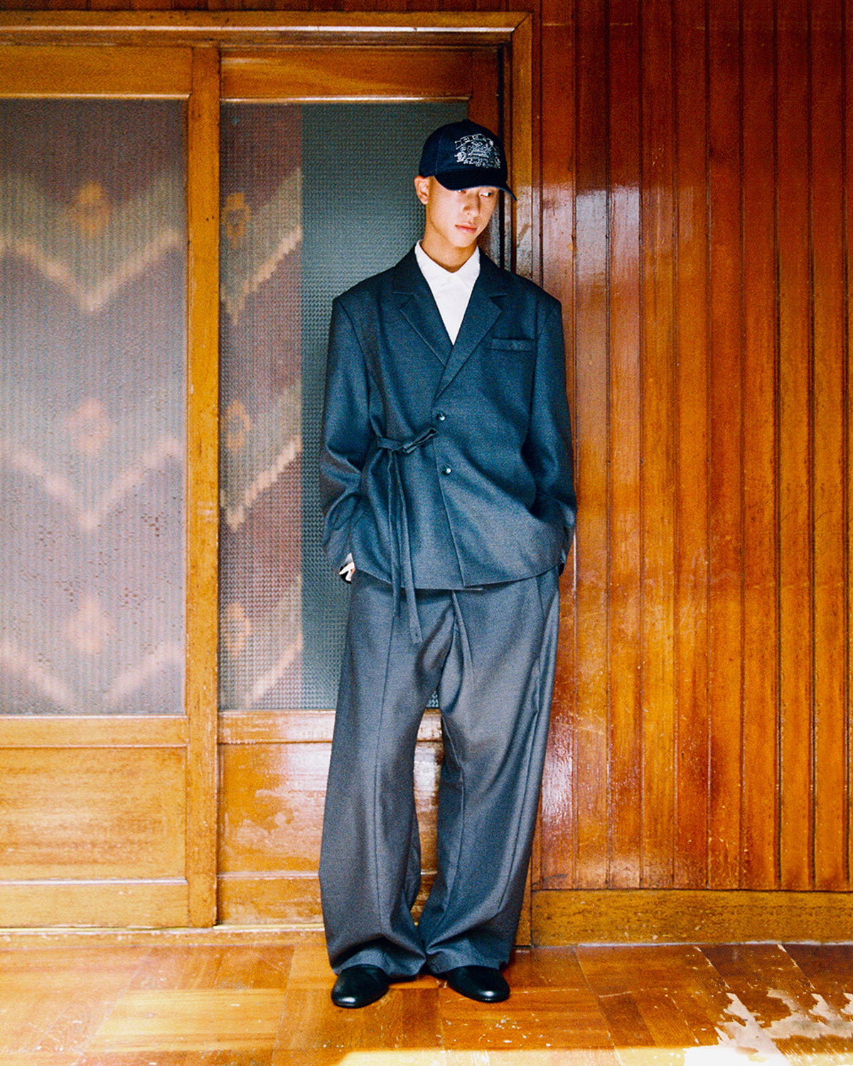 A model in a black cap wears the Sundae School CHARCOAL DOUBLE DUTY BLAZER tied at the waist with matching CHARCOAL PLEATED HANBOK SLACKS, by a wooden door.