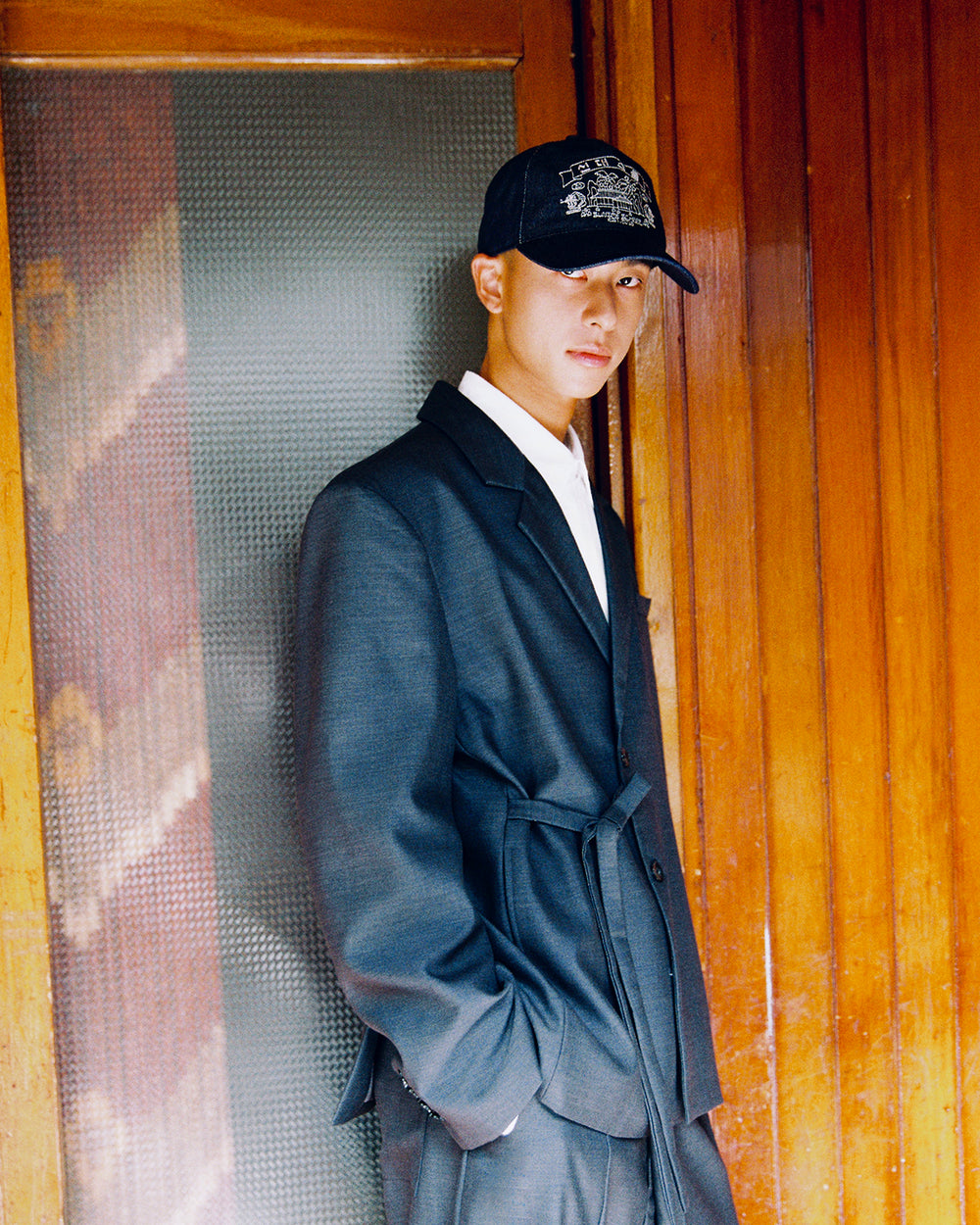 A model leans against a wooden door wearing the Sundae School CHARCOAL DOUBLE DUTY BLAZER over a white shirt with matching charcoal slacks.