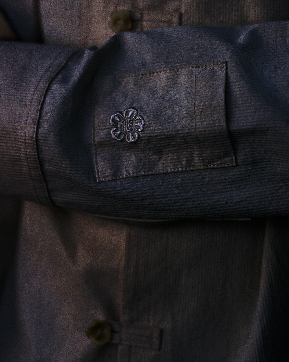 A close-up of a person in the Sundae School BLACK CHINA COLLAR KNOT SHACKET, featuring folded arms, a square patch with embroidered florals on the sleeve, visible seams in corded cotton twill, and large dark buttons.