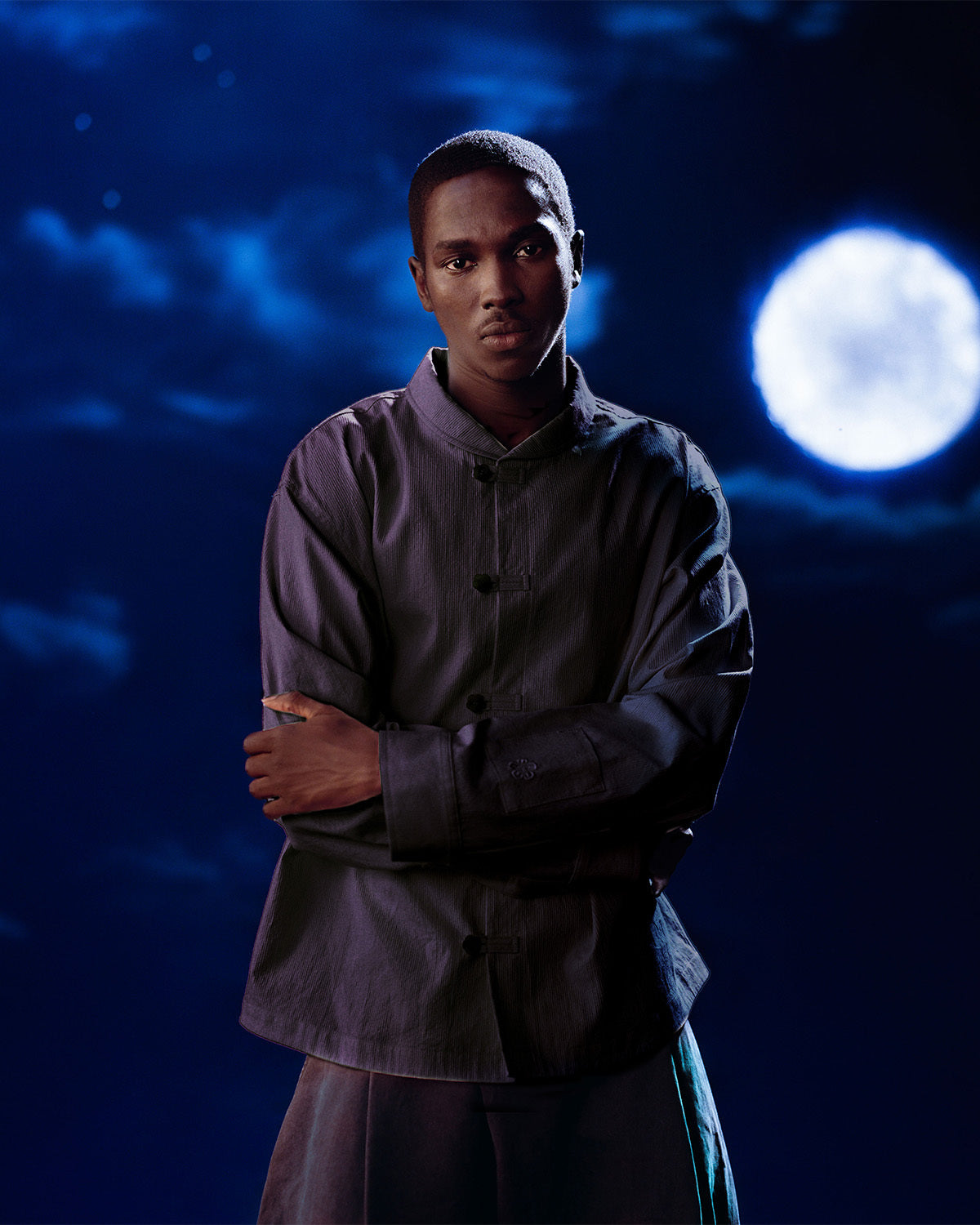 A person stands with arms crossed, wearing the BLACK CHINA COLLAR KNOT SHACKET by Sundae School—a dark corded cotton twill button-up—against a night sky with clouds and a bright full moon.