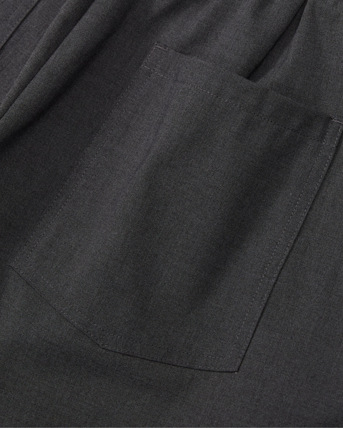 Close-up of dark gray fabric with a rectangular patch pocket and visible stitching, on Sundae School's FLARE SKIRT PANTS. The smooth, subtly textured material is perfect for summer wear.