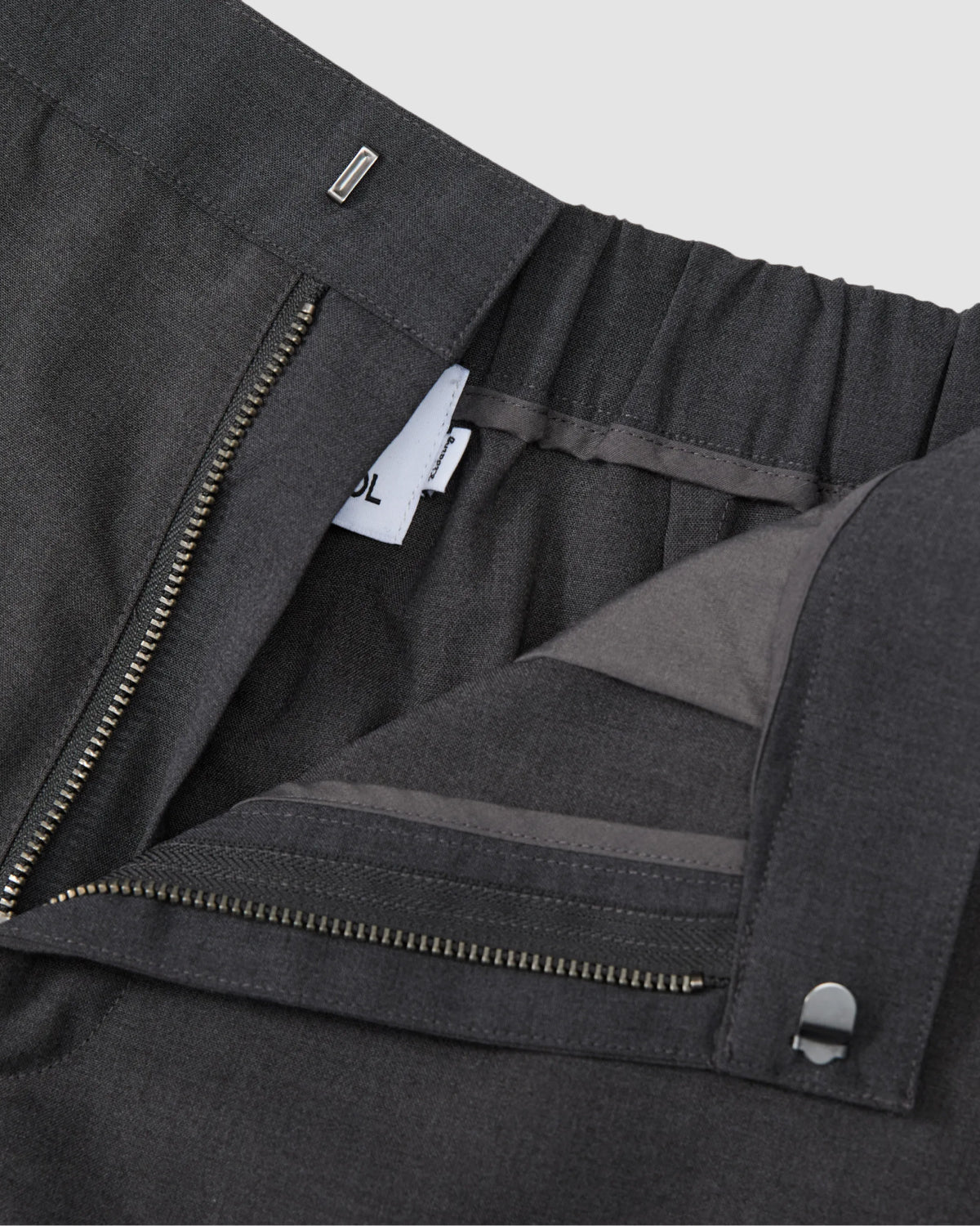 Close-up of Sundae School FLARE SKIRT PANTS in dark gray summer wool, featuring an open zipper fly, elastic waistband, visible inner fabric layers, and a partially visible white tag with black text inside.