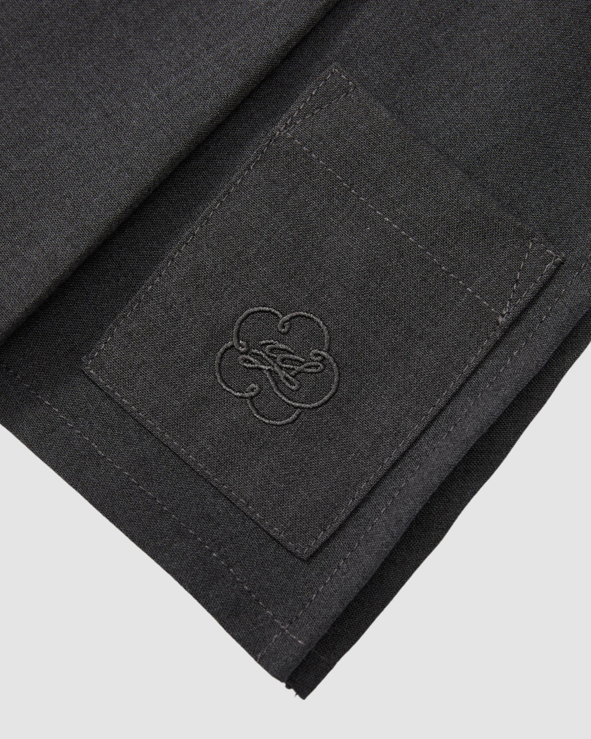 Close-up of dark fabric on Sundae School's FLARE SKIRT PANTS featuring a stitched rectangular pocket with an embroidered clover design and cursive text, neatly finished—ideal for pairing with summer wool trousers.