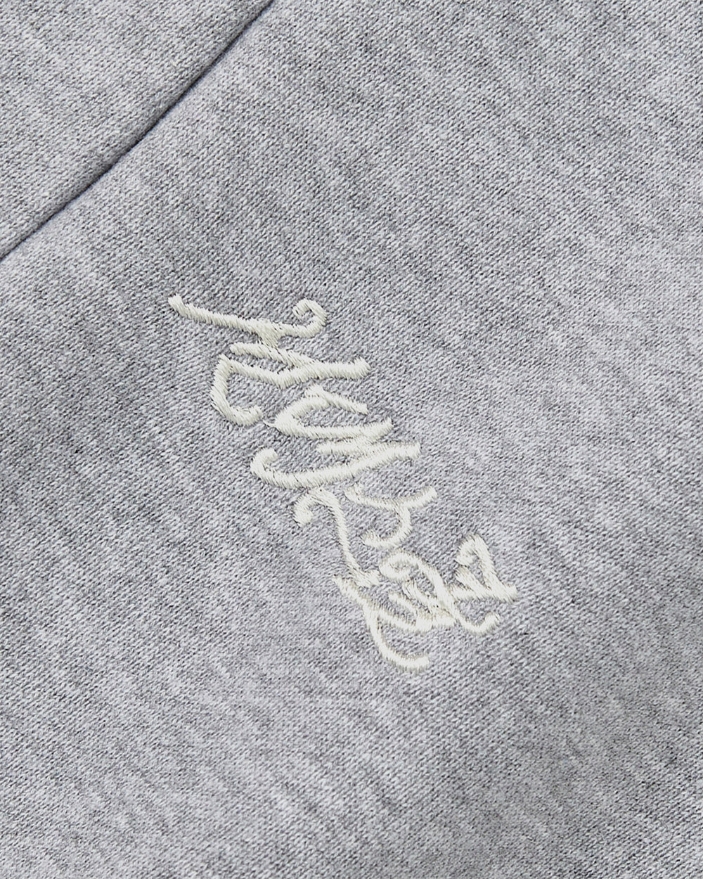 A close-up of heather grey fabric from Sundae School’s HEATHER GREY WRAP SWEATBAJI, featuring delicate white Korean embroidery in cursive. Inspired by hanbok wrap designs, the decorative stitching lends a traditional accent, even as the script stays hard to decipher.