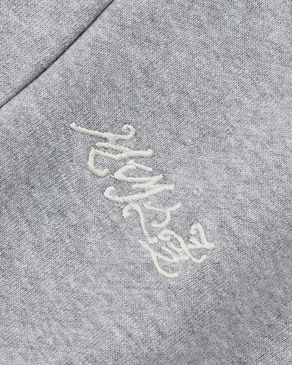 A close-up of heather grey fabric from Sundae School’s HEATHER GREY WRAP SWEATBAJI, featuring delicate white Korean embroidery in cursive. Inspired by hanbok wrap designs, the decorative stitching lends a traditional accent, even as the script stays hard to decipher.