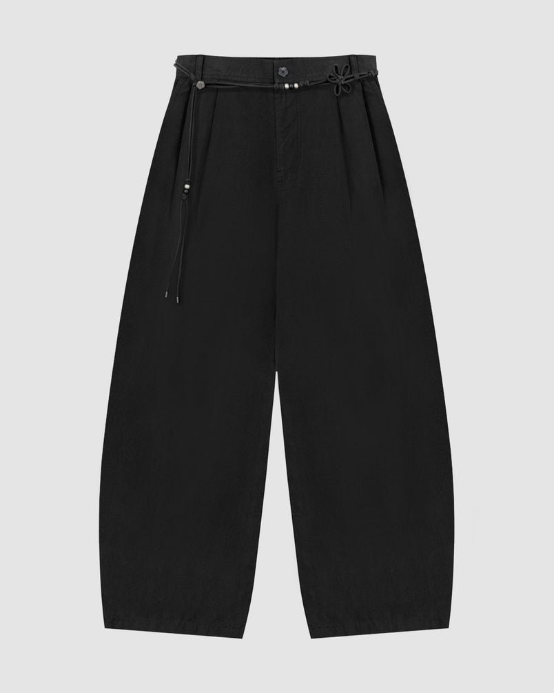 The BLACK FLOWER NORIGAE LINEN TROUSERS by Sundae School feature a wide-leg, balloon silhouette in black cotton linen, pleated at the waist and accented with a norigae belt adorned with bead details. The minimalist design is showcased against a plain light gray background.