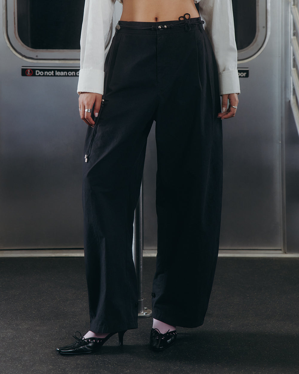 A person stands in a subway car wearing Sundae School's BLACK FLOWER NORIGAE LINEN TROUSERS paired with a white shirt, black heels, and pale socks. Their head is out of frame, and a sign behind them reads, "Do not lean on door.