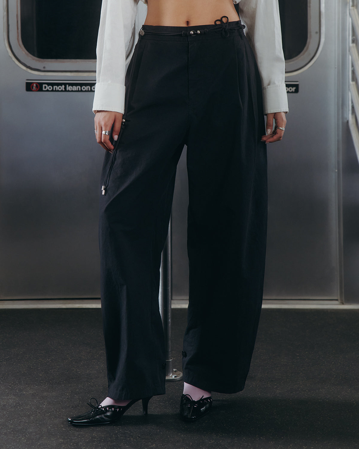 A person stands in a subway car wearing Sundae School's BLACK FLOWER NORIGAE LINEN TROUSERS paired with a white shirt, black heels, and pale socks. Their head is out of frame, and a sign behind them reads, "Do not lean on door.