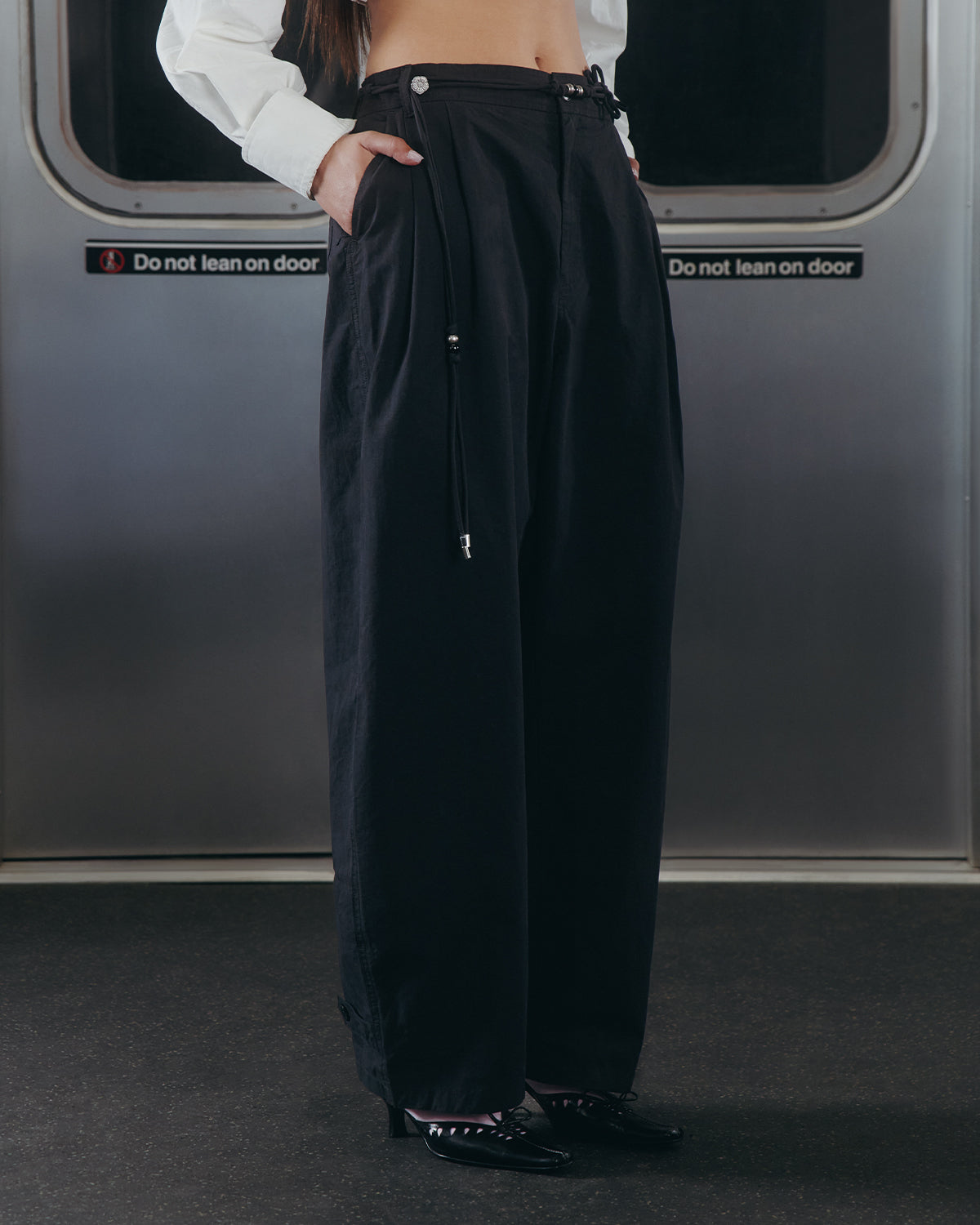 A person wearing Sundae School's BLACK FLOWER NORIGAE LINEN TROUSERS and black shoes stands in front of a subway door with 