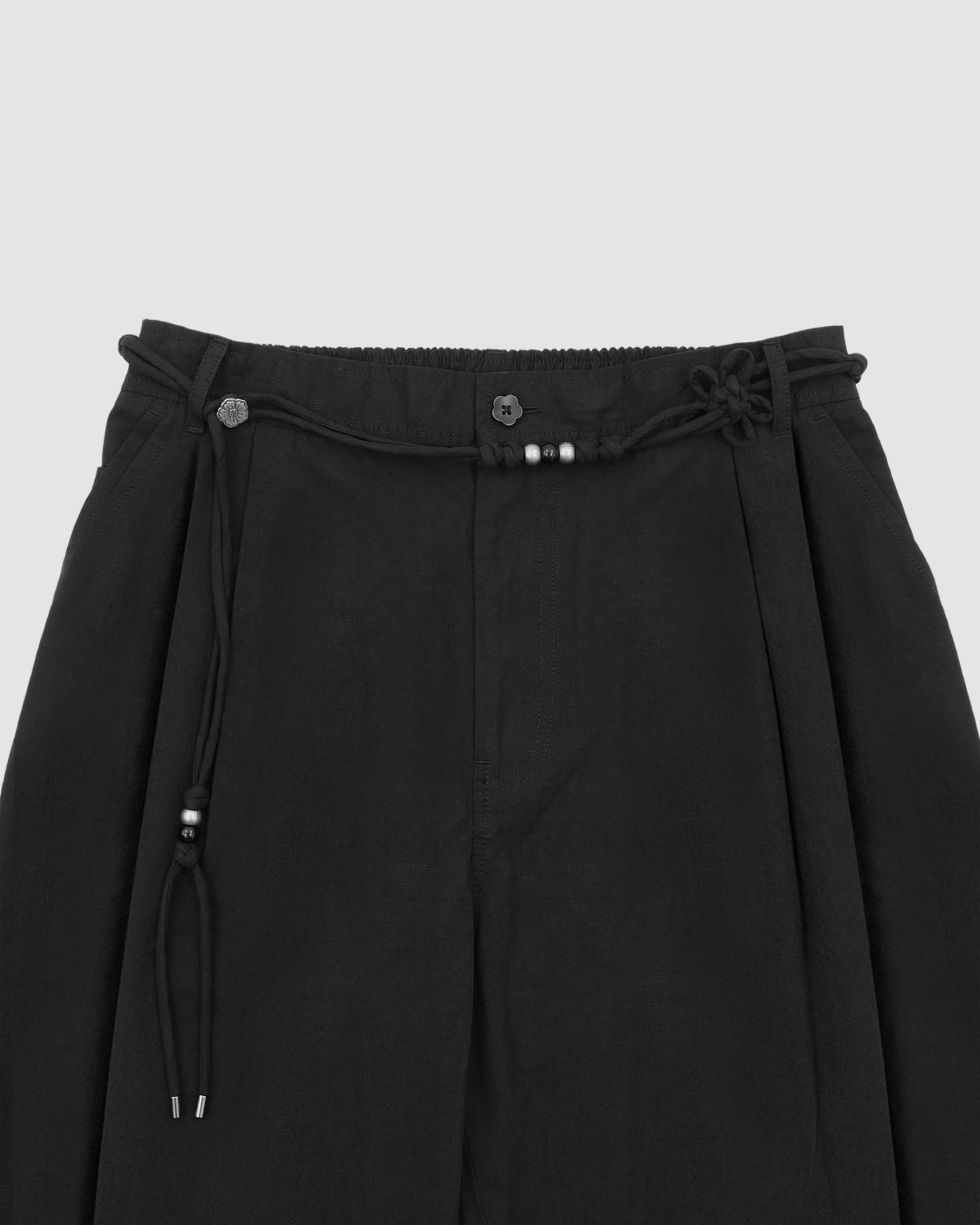 Sundae School’s BLACK FLOWER NORIGAE LINEN TROUSERS, crafted from pleated black cotton-linen and detailed with a silver-beaded norigae belt and subtle stash pocket, are displayed against a light gray background.