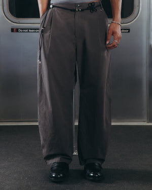 A person wearing Sundae School’s CHARCOAL FLOWER NORIGAE LINEN TROUSERS—loose, dark gray pants with a black drawstring—paired with black shoes is standing on a subway train, with only the lower half of their body visible.