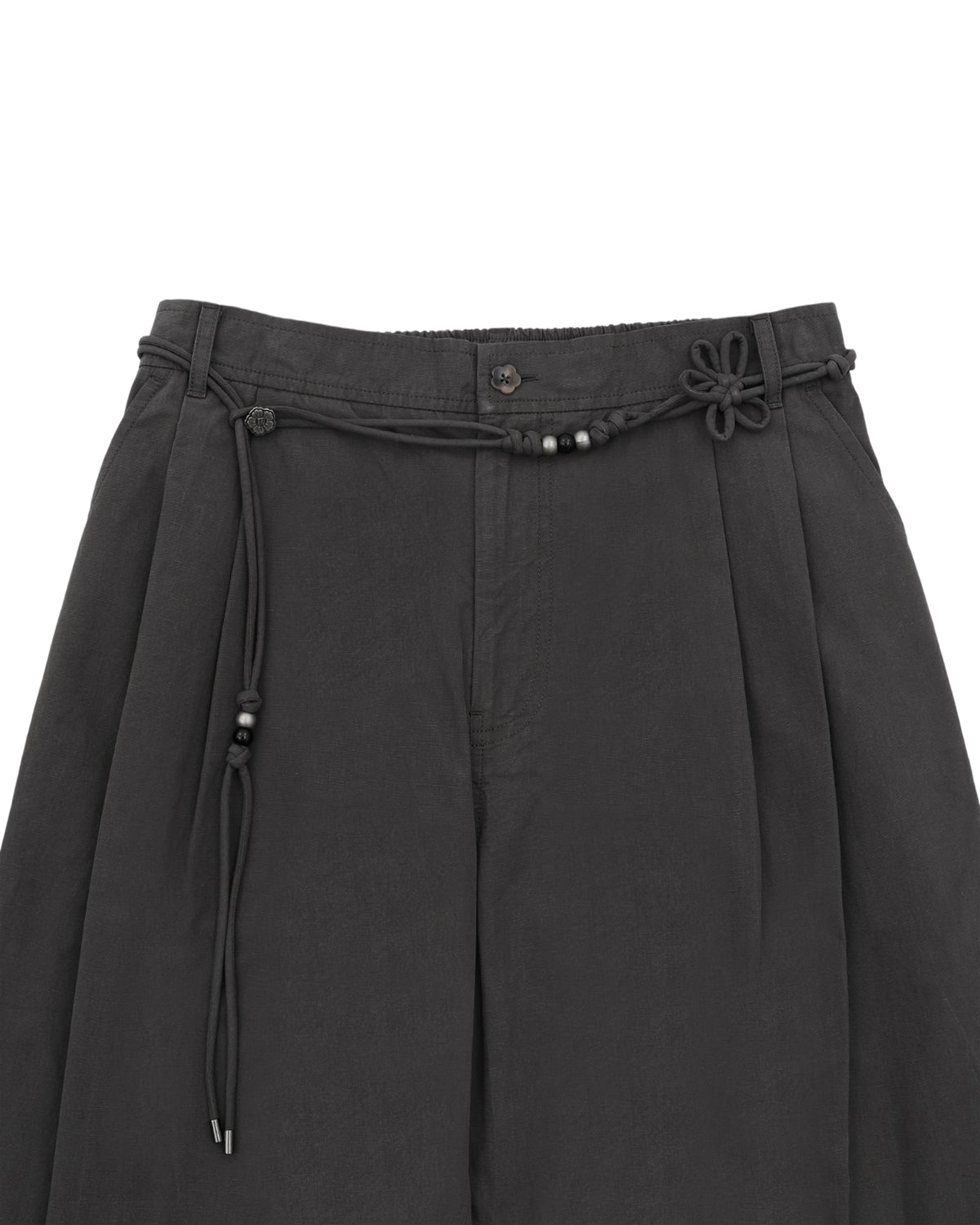 The CHARCOAL FLOWER NORIGAE LINEN TROUSERS from Sundae School are crafted in a dark gray cotton-linen blend and feature elegant pleats. A decorative black norigae belt with beaded details and a flower-shaped knot accents the right side, while the modern balloon leg silhouette is complemented by practical belt loops and a button closure at the waistband.