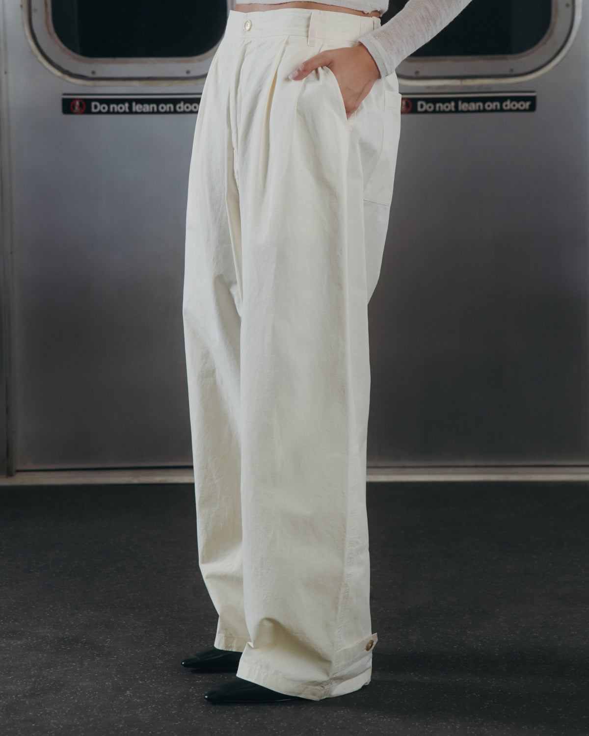 A person wearing Sundae School’s IVORY FLOWER NORIGAE LINEN TROUSERS stands in a subway car with hands in pockets, framed by metal subway doors featuring "Do not lean on door" signs.