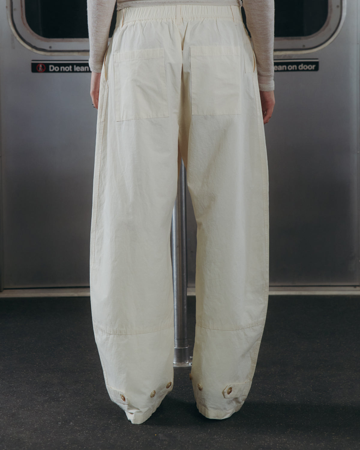A person wearing Sundae School's IVORY FLOWER NORIGAE LINEN TROUSERS stands on public transportation, facing away. The background features a metal door with a sign that reads, 