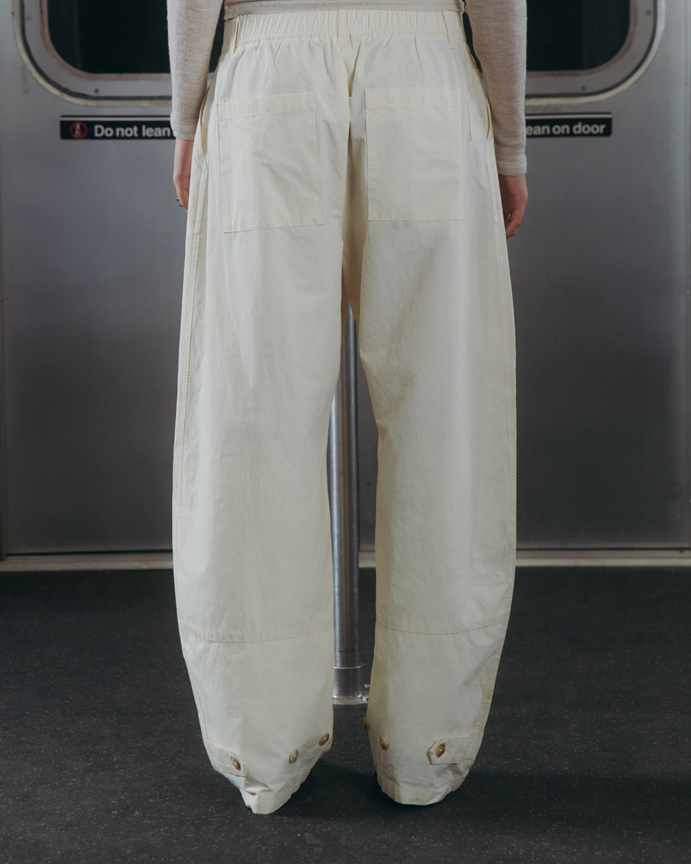 A person wearing Sundae School's IVORY FLOWER NORIGAE LINEN TROUSERS stands on public transportation, facing away. The background features a metal door with a sign that reads, "Do not lean on door.