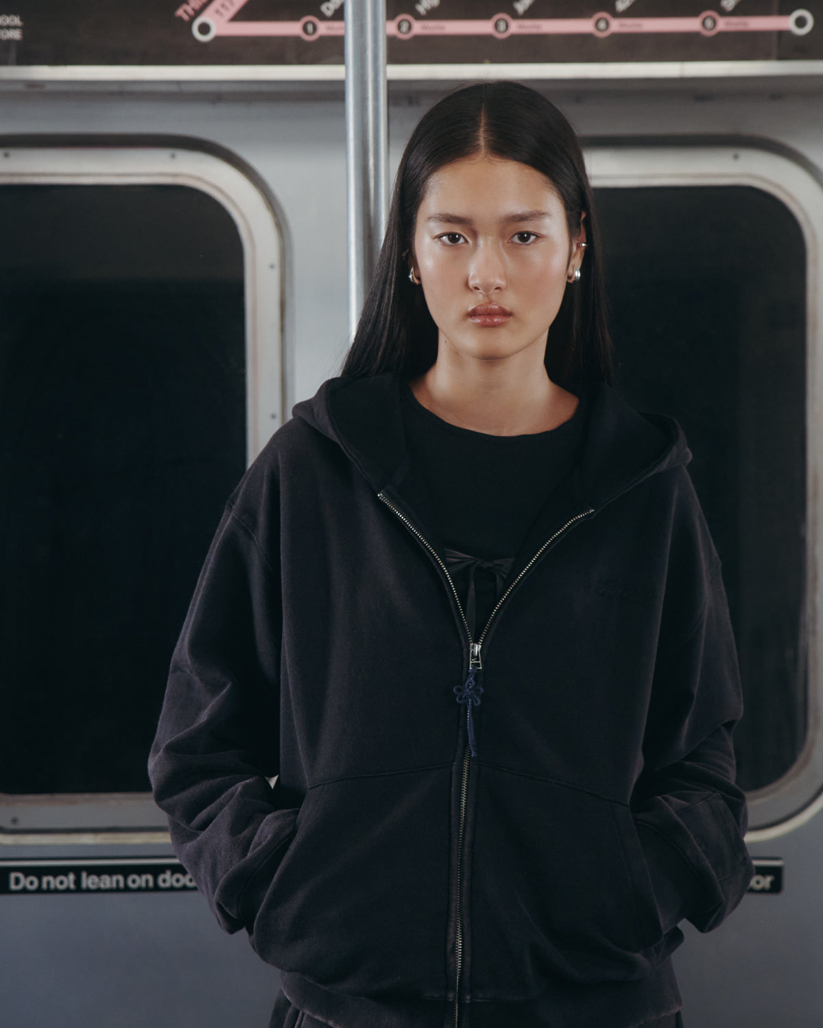 A woman with long dark hair stands in a subway car, wearing the Sundae School BLACK WASH FLOWER KNOT ZIP UP hoodie with her hands in her pockets. The hoodie features a Maehwa knot zipper pull and subtle Hangul Sundae School embroidery. A subway route map and warning sign are visible in the background.