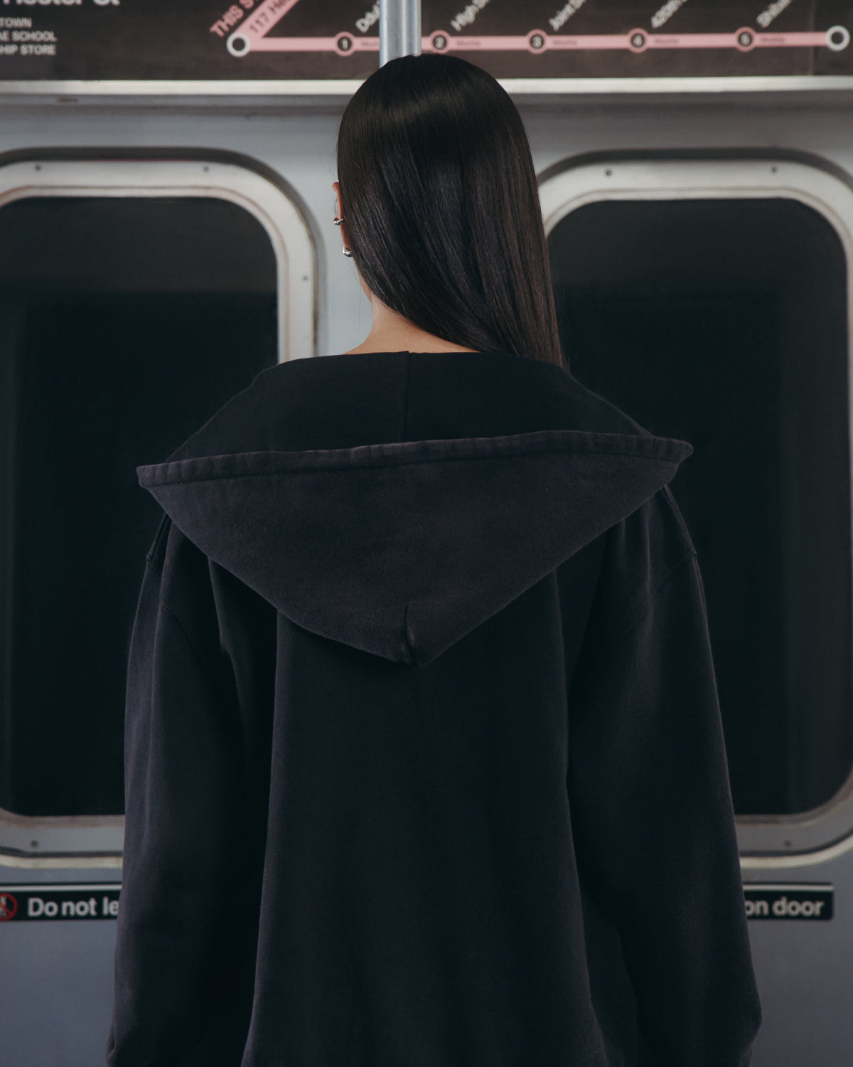 A person with long, straight hair stands inside a subway train, facing away from the camera and gazing out the window. They are wearing Sundae School's BLACK WASH FLOWER KNOT ZIP UP hoodie, distinguished by a Maehwa knot zipper pull and subtle Hangul Sundae School embroidery on the back. A subway map and safety sign can be seen in the background.