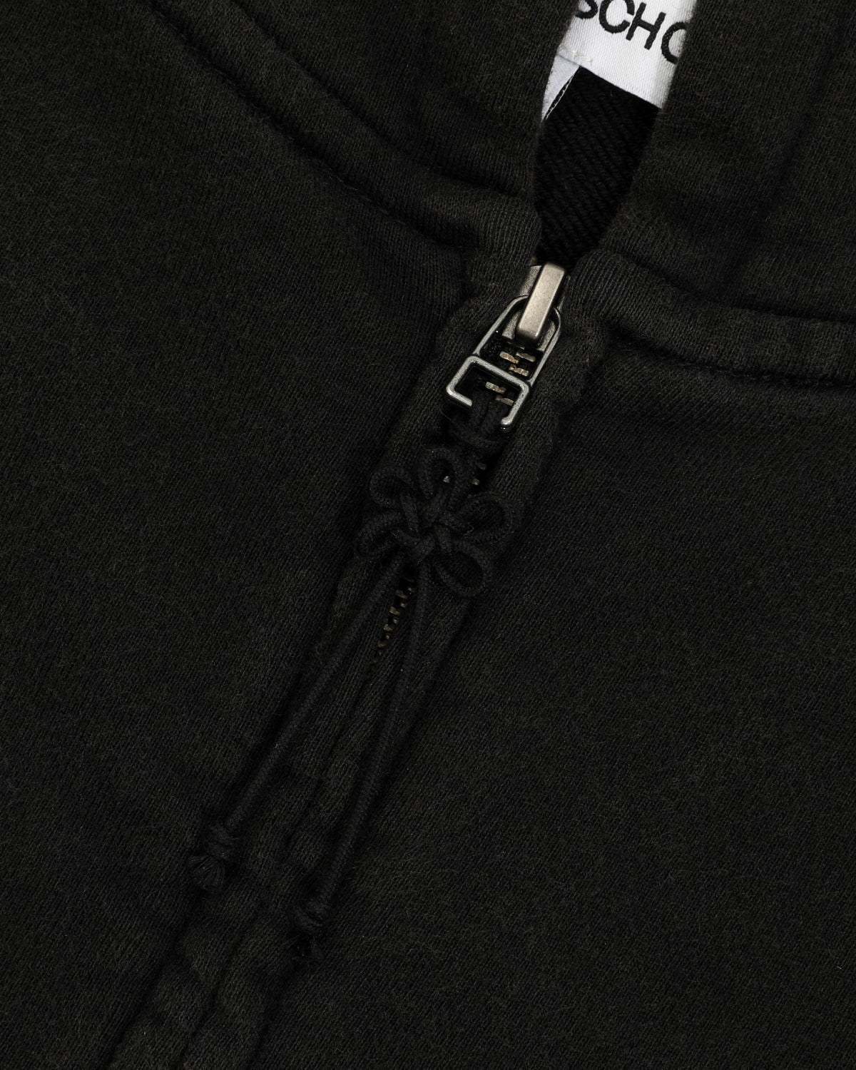 Close-up of the BLACK WASH FLOWER KNOT ZIP UP by Sundae School, highlighting the black cotton fabric, zipper with a Maehwa knot pull, and drawstrings. The top of the zipper is partially open, revealing a glimpse of a white tag with black text inside the collar.