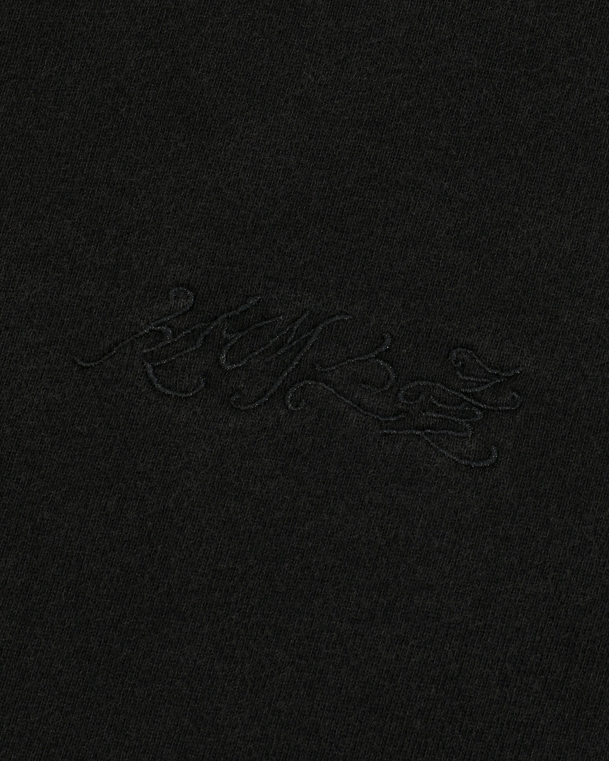 A close-up of black fabric featuring subtle, decorative black embroidered script that blends seamlessly with the material highlights the unique Hangul Sundae School embroidery on the BLACK WASH FLOWER KNOT ZIP UP hoodie by Sundae School.