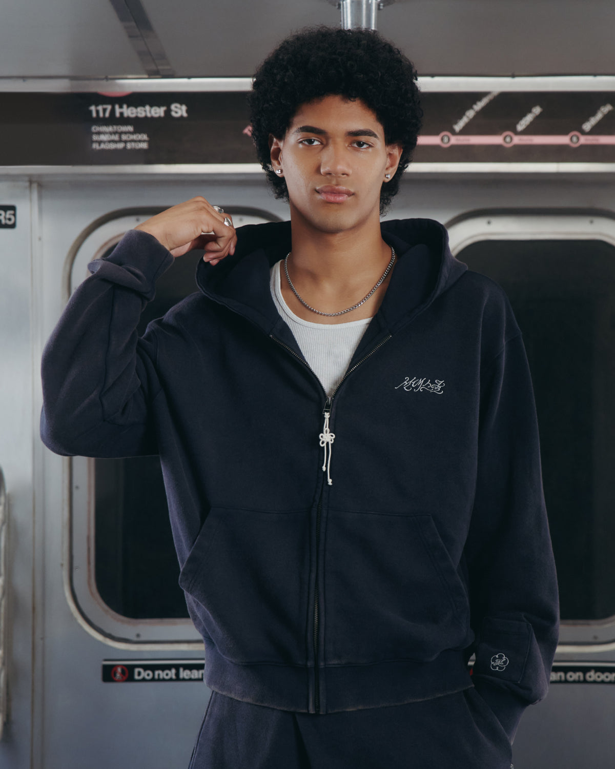A young man with curly hair wearing the Sundae School NAVY WASH FLOWER KNOT ZIP UP hoodie and a white shirt stands in a subway train, holding his hood, with the 