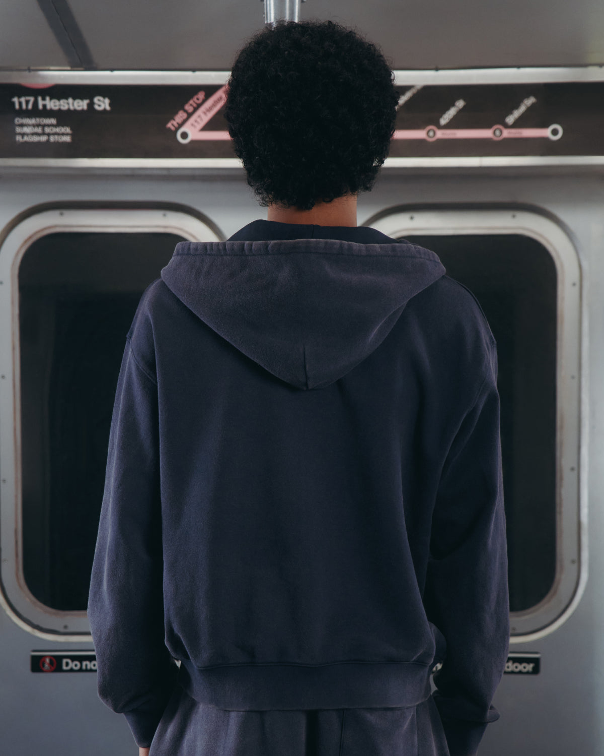 A person with short curly hair, wearing the Sundae School NAVY WASH FLOWER KNOT ZIP UP, stands facing away inside a subway train. A subway map and a 