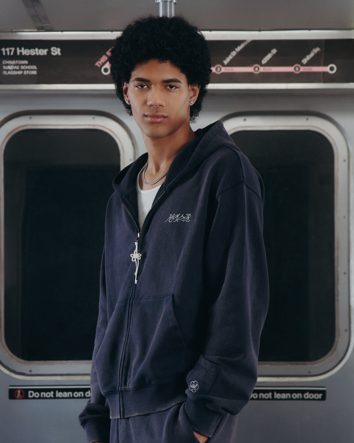 A young person with curly hair stands inside a subway train, in front of doors and a sign that reads 117 Hester St., wearing the NAVY WASH FLOWER KNOT ZIP UP from Sundae School. With a neutral expression, they look into the camera, effortlessly capturing the cool street vibe of Hangul Sundae School.